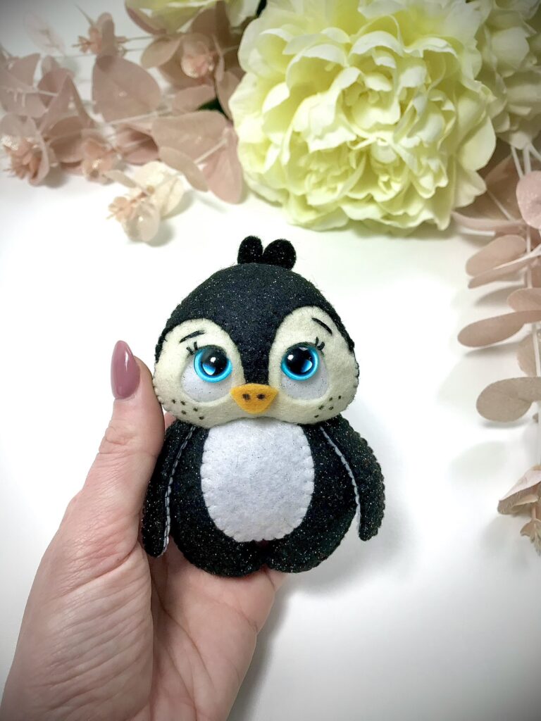 Person holding a handmade felt penguin ornament with large blue eyes, against a soft background of white and pale green flowers.