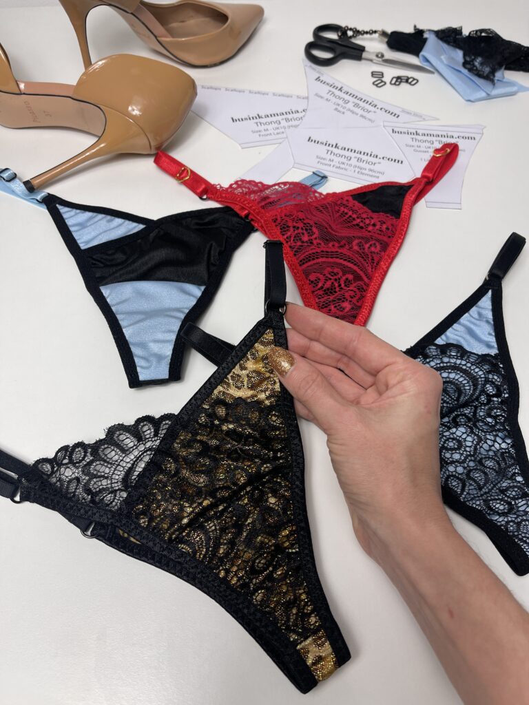Hand holding black and gold lace bralette beside red and black designs, sewing patterns, and high heel shoe on table.