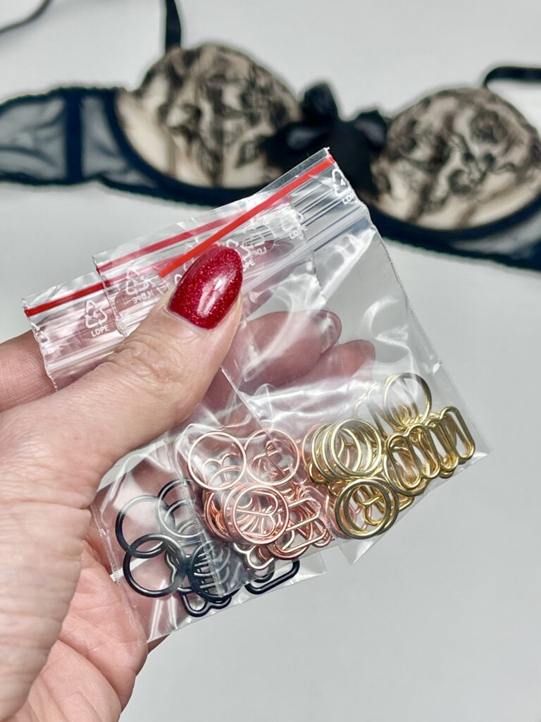 Hand holding bags of lingerie hardware in gold, rose gold, and black, with lace bra in background.