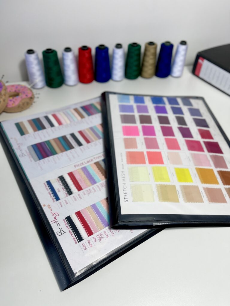 Colour charts and thread sample cards on a white table, with colourful spools of thread displayed in the background
