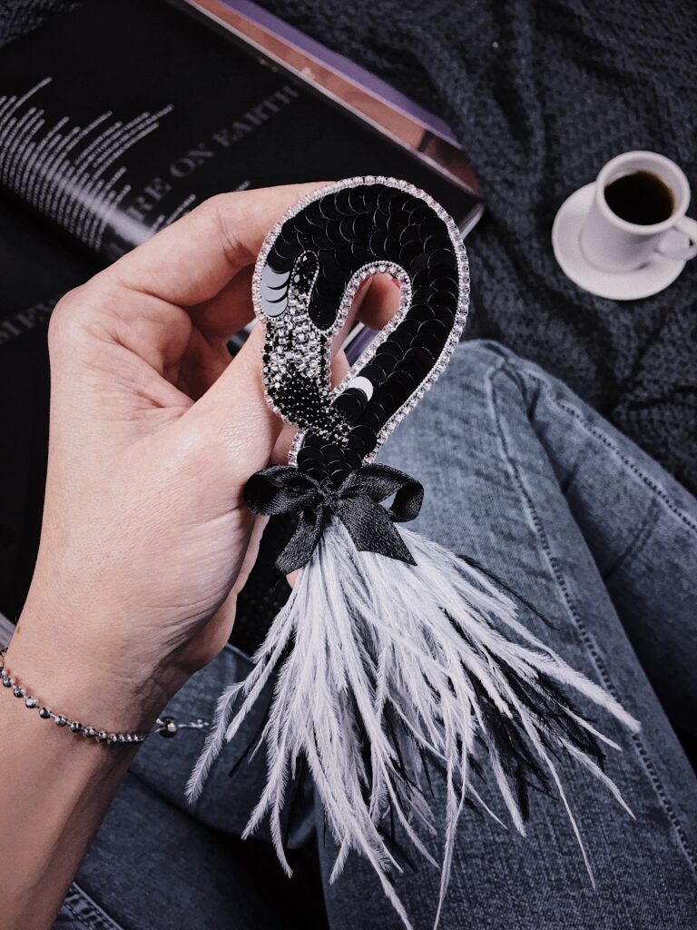 Hand holding a black sequin embroidered flamingo brooch with a bow and feather fringe, made on hard felt.