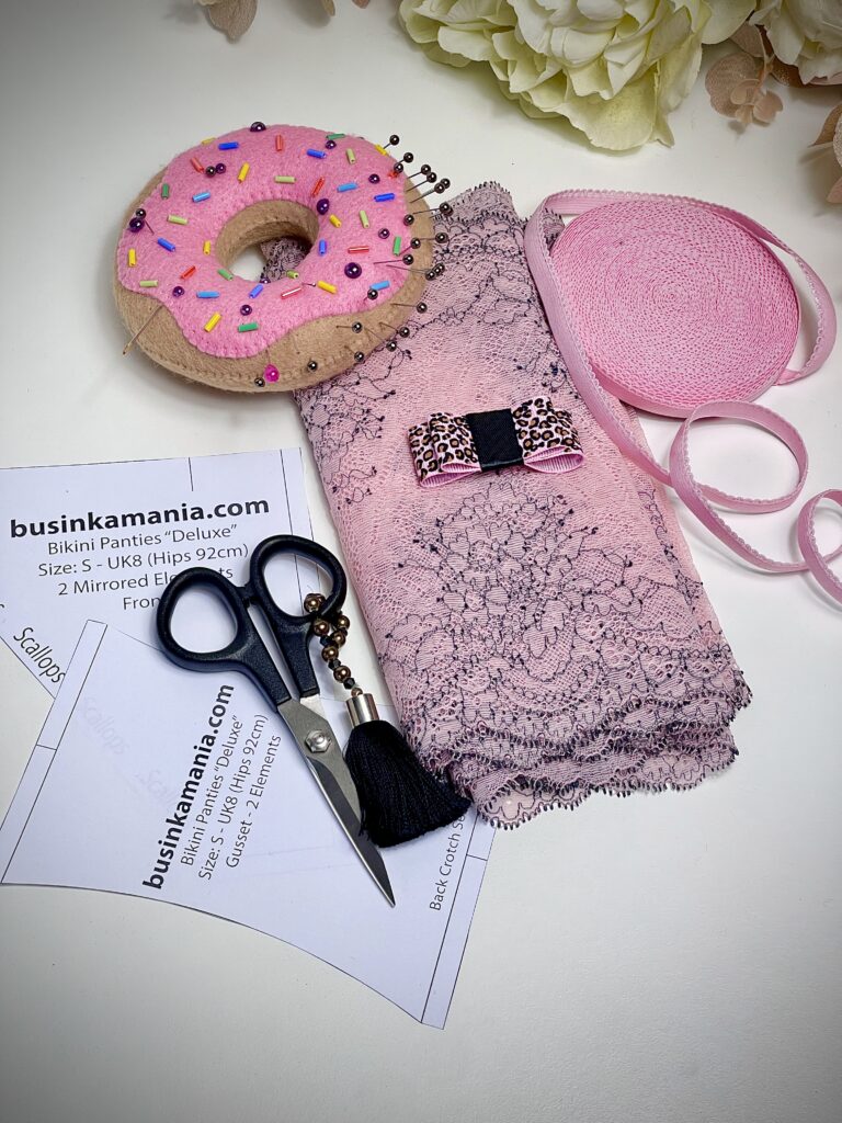 Pink lace fabric with lingerie sewing supplies, paper pattern, ribbons, scissors, and pin cushion, ready for DIY underwear projectD