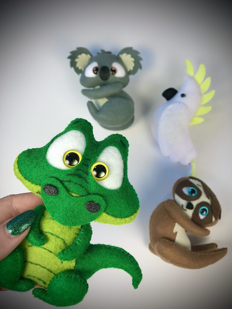 Hand holding a green felt crocodile plush toy, with three more handmade felt animals (koala, cockatoo, and sloth) displayed in the background.