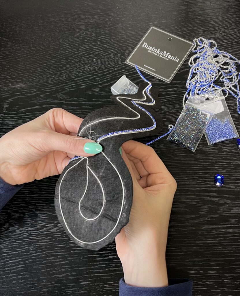Close-up of hands sewing bead embroidery on black hard felt, with seed beads, crystals, and jewellery-making supplies on a black table.
