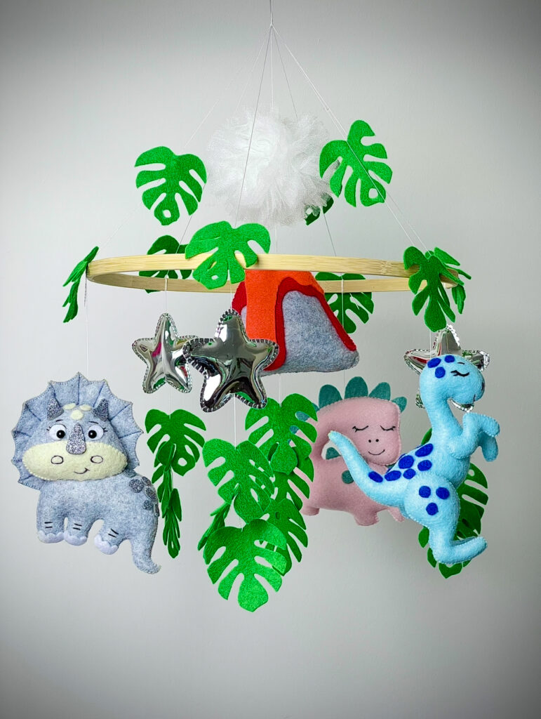 Handmade felt dinosaur-themed baby cot mobile with green leaves, silver stars, volcano, and colourful dinosaur plushies.