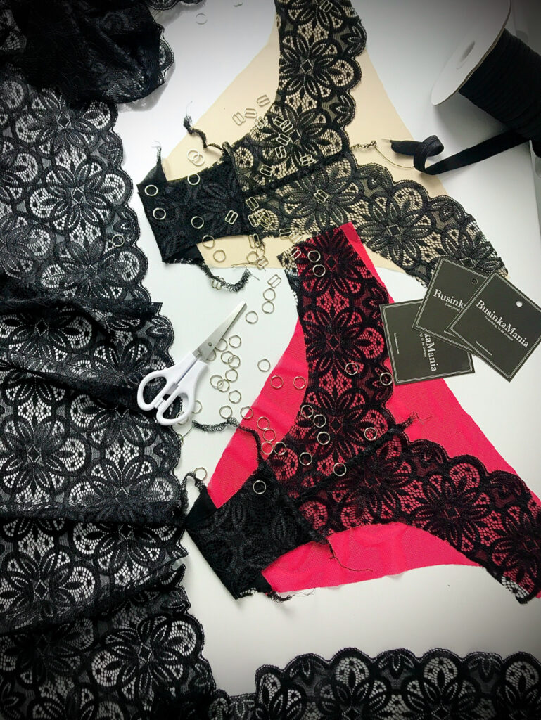 Black lace, pink lingerie, silver rings, white scissors, and branded tags lay ready for a handmade lingerie project.
