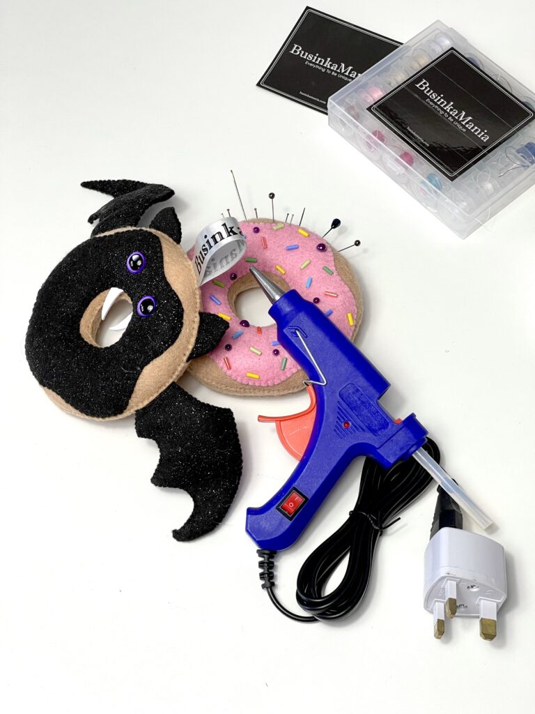 Blue hot glue gun next to two handmade felt donut toys, one decorated as a bat and the other as a pink sprinkle donut with pins inserted.