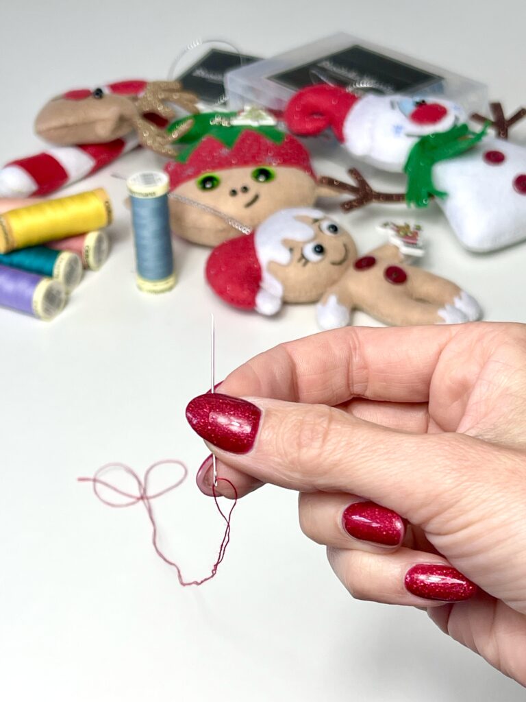 Hand holding a threaded needle with red thread, surrounded by colourful sewing threads and handmade felt Christmas ornaments like gingerbread and snowman.
