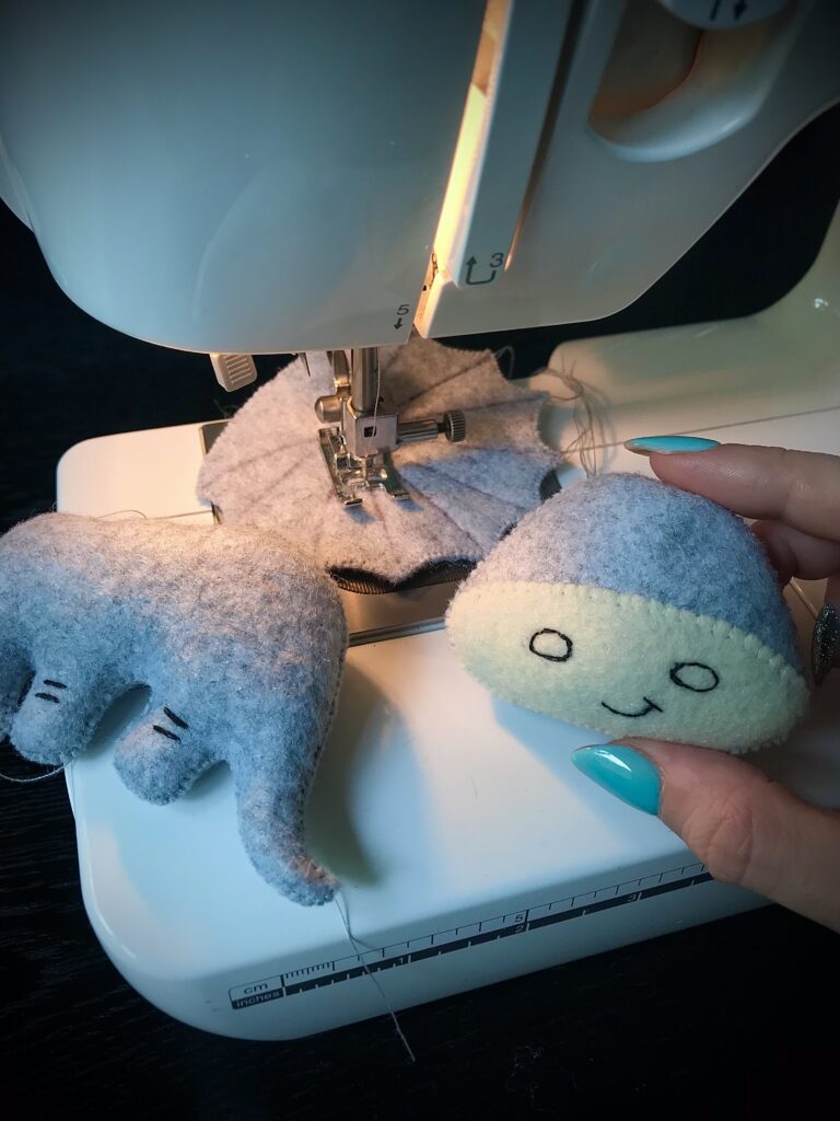 Grey felt dinosaur toy in progress on sewing machine, with a finished grey and cream dinosaur held by hand.