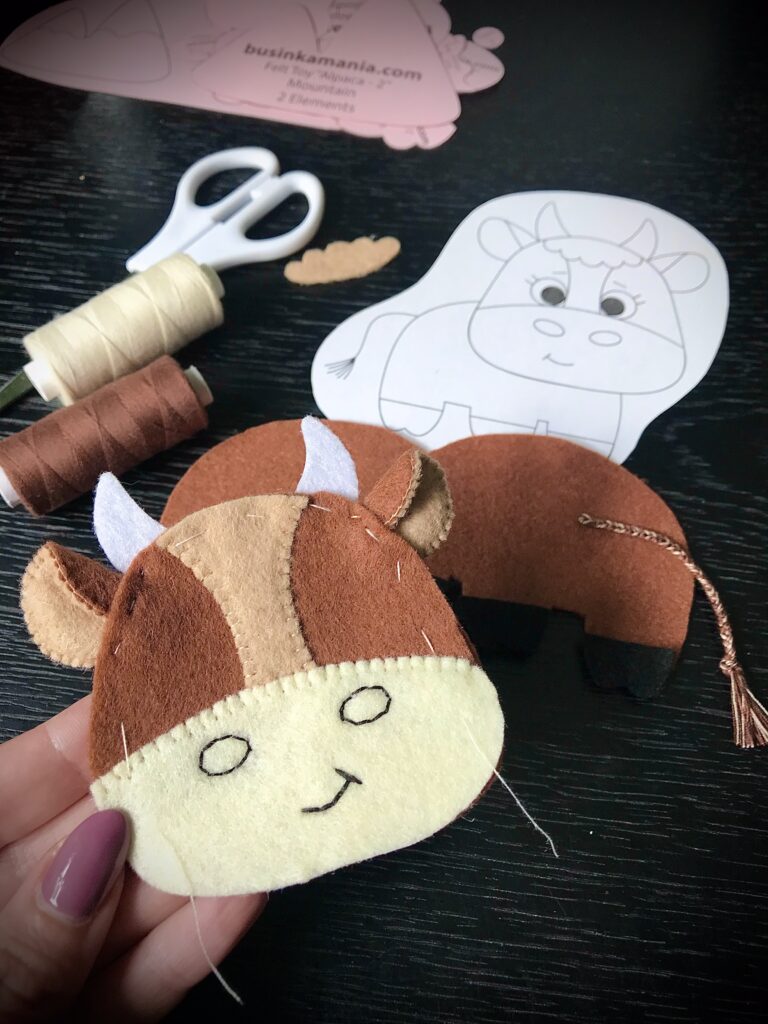 Felt cow toy in progress using whip stitch, with sewing supplies and a printed cow pattern in the background.