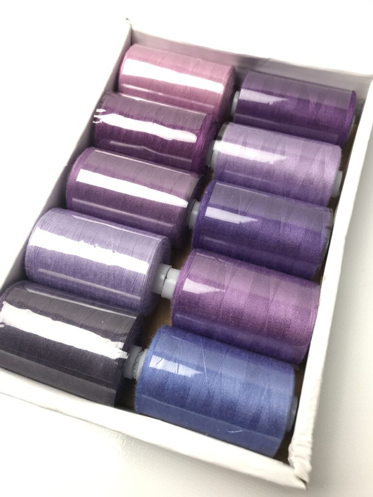 Ten spools of sewing thread in various shades of purple, neatly arranged in a white box with a shiny finish.