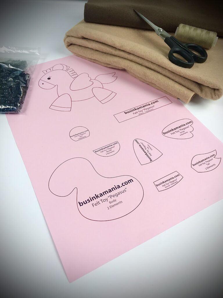 Printed felt Pegasus sewing pattern on pink paper with beige felt, scissors, and toy eyes ready for crafting.