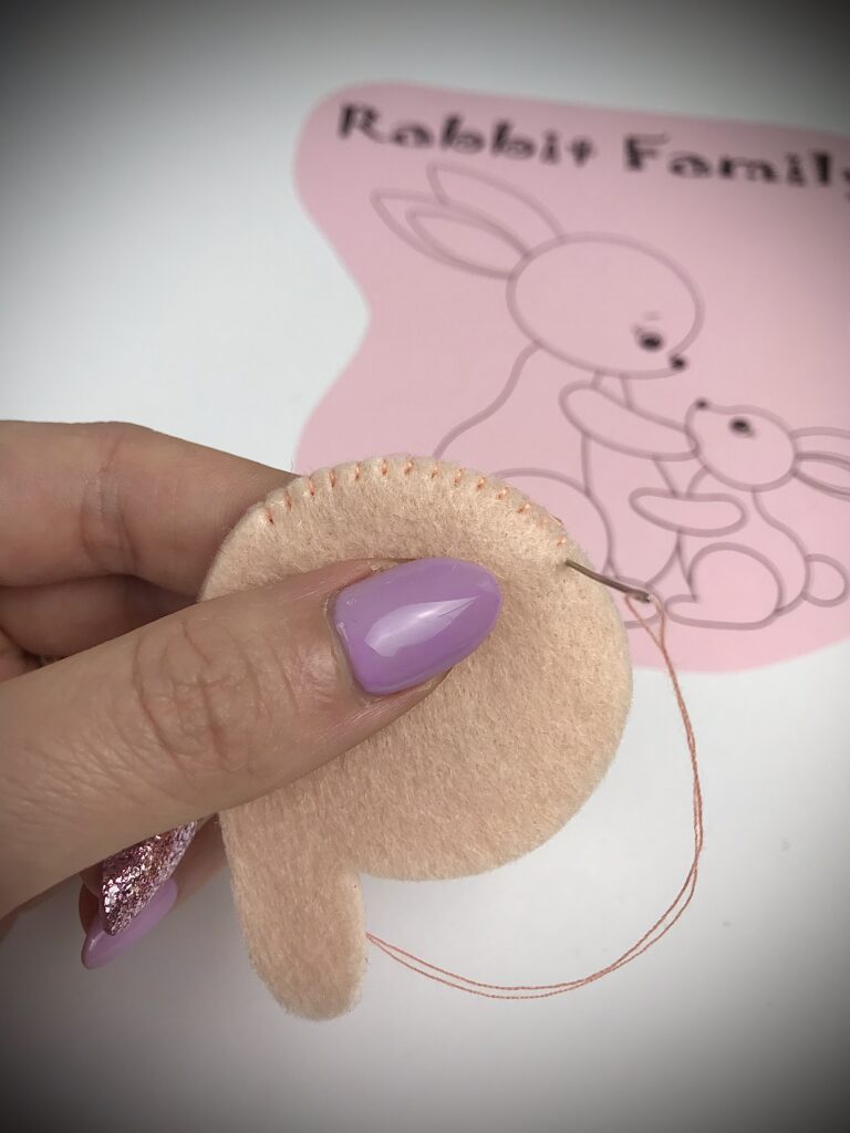 Hand sewing peach felt bunny part with blanket stitch, in front of a pink bunny family pattern background.