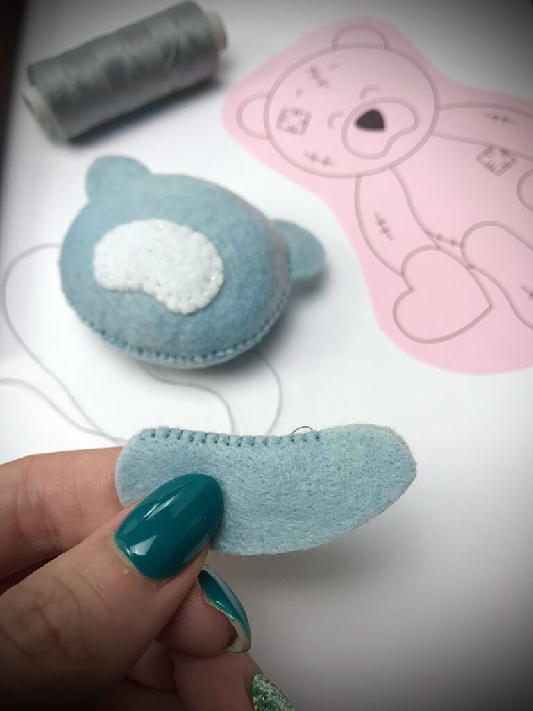 Sewing a blue felt teddy bear part by hand, with bear head and printed pattern visible in the background.