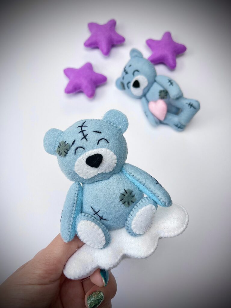 Blue felt teddy bear toy with stitched details, held on a cloud, with stars and another bear plush in the background.