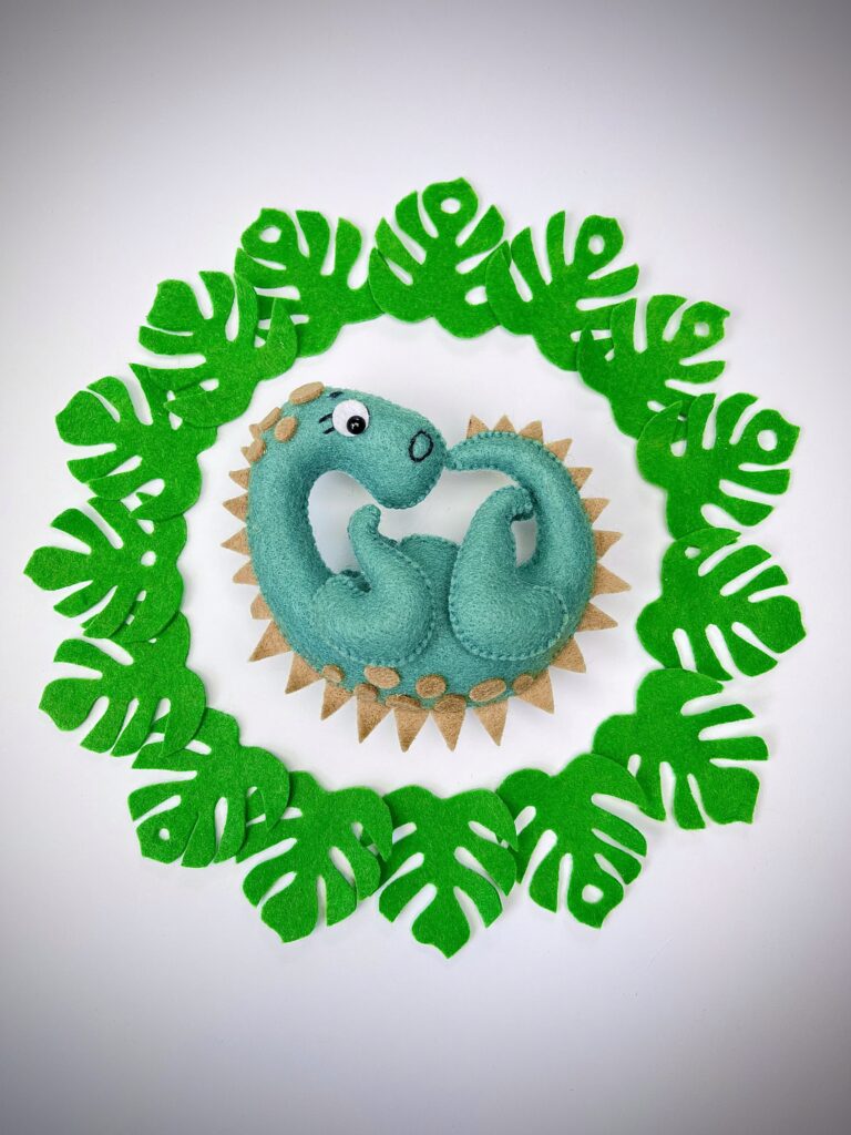 Green felt dinosaur plush surrounded by a circular frame of bright green felt jungle leaves on a white background.