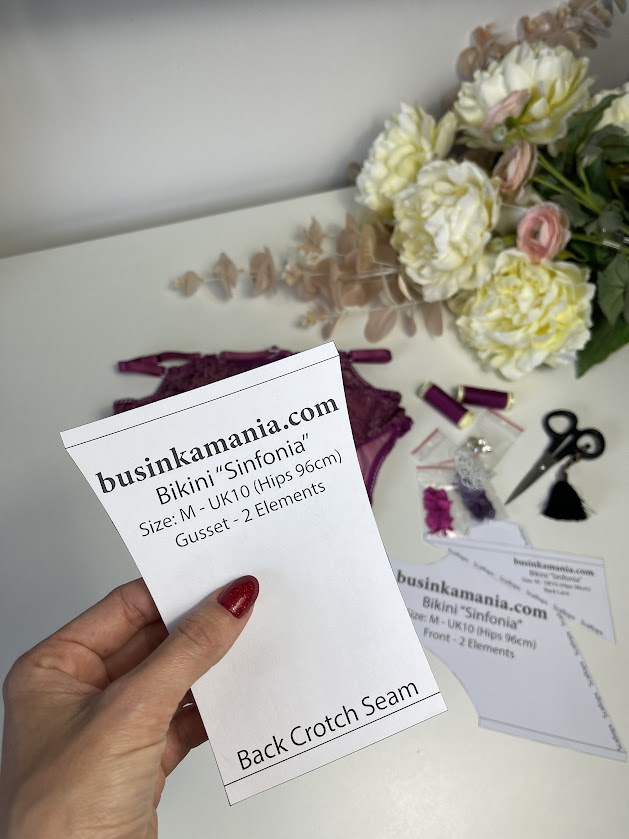 Hand holding lingerie sewing pattern piece with gusset detail, surrounded by sewing tools and lace fabric on a table.