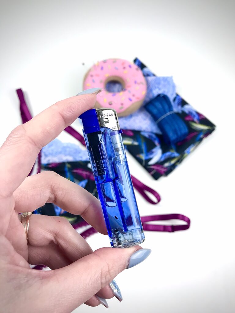 Hand holding blue lighter for sealing thread ends with sewing supplies, fabric and elastic in lingerie finishing tutorial.