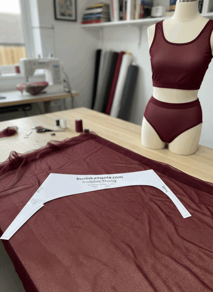 Invisible thong sewing pattern placed on burgundy stretch mesh fabric beside mannequin in matching handmade lingerie set.