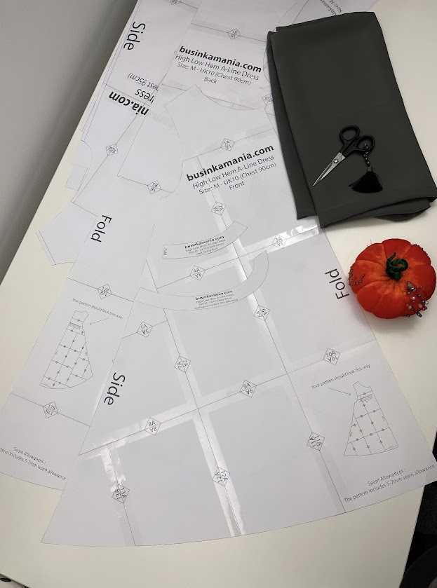 Large BusinkaMania dress sewing pattern assembled on table with folded grey fabric, scissors, and red tomato pin cushion.