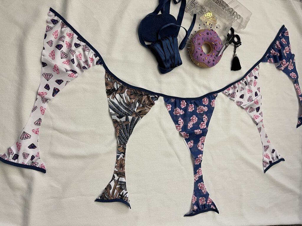 Five handmade thong panties with fold-over elastic sewn on edges, displayed with sewing tools for Step 3 elastic tutorial.