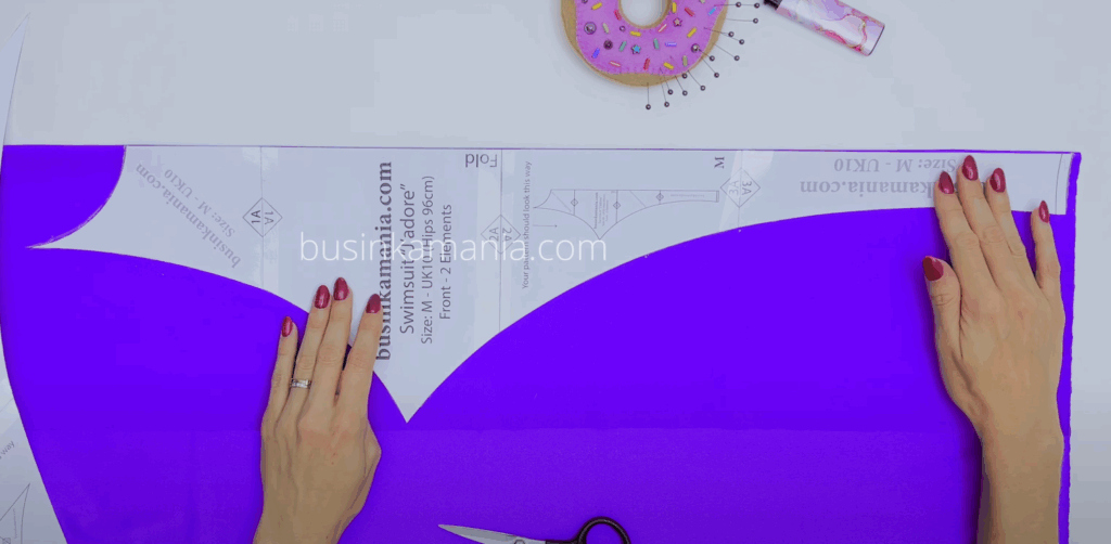 Hands placing Swimsuit Jadore pattern on folded purple fabric, preparing to trace and cut pieces using chalk.