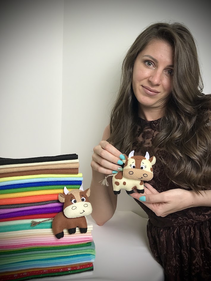 Woman holding felt cow plush with braided thread tail and tassel beside stacked colourful soft felt sheets.