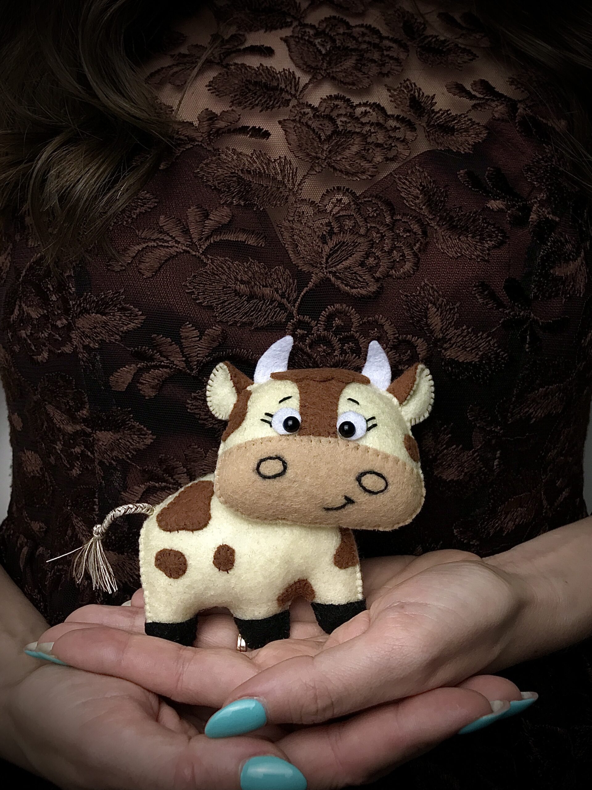 Hand holding felt cow plush with braided tassel tail detail for decorative felt animal sewing project.