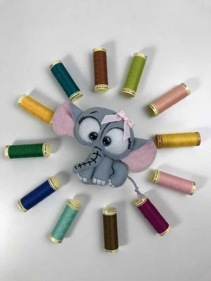 Grey felt elephant plush with chain stitch facial details surrounded by colourful thread spools for felt toy tutorial