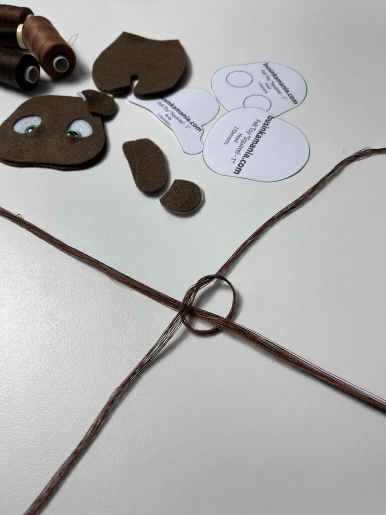 Threads crossed in T shape and knotted for braided tail, beside felt cow pattern pieces and polyester thread spools.