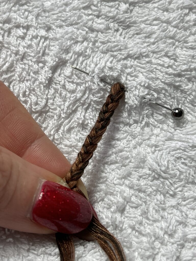 Braiding polyester threads pinned in place on fabric to form decorative tassel tail for felt plush animal.