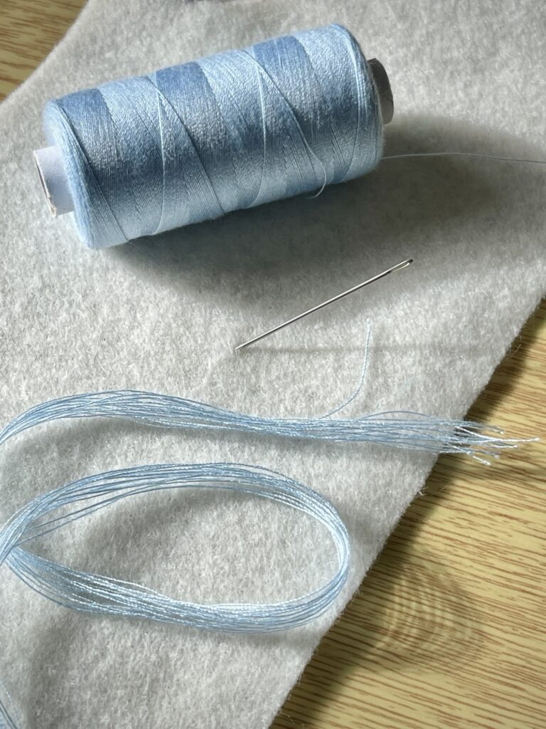 Light blue sewing thread, needle and folded strands on soft felt, materials for making tassels on craft project.