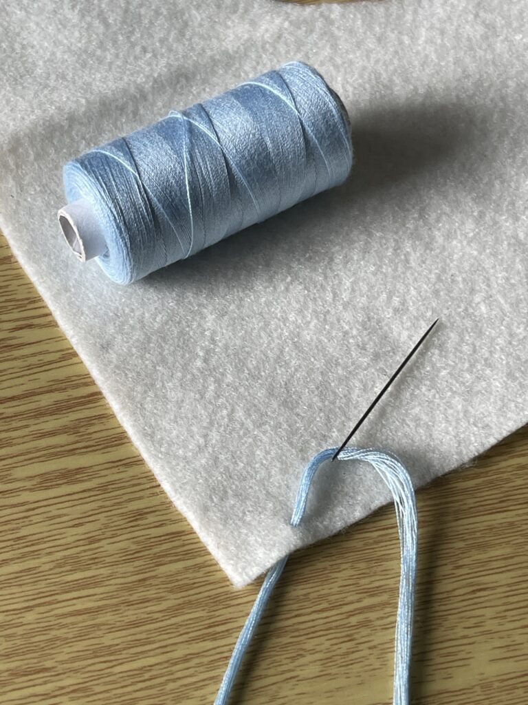 Folded light blue threads on needle over soft felt, ready to place tassel on handmade craft project.