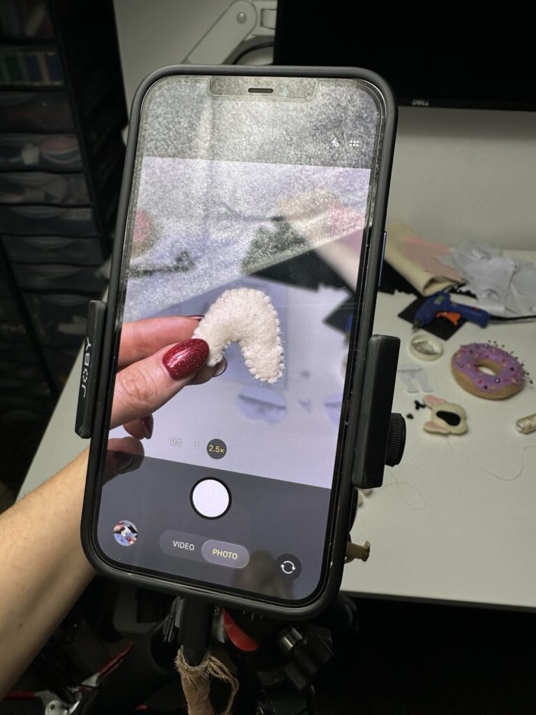 Recording felt plushie sewing process on phone while holding stitched piece, showing behind the scenes of handmade French bulldog toy