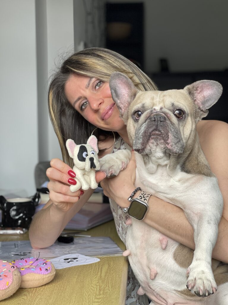 Woman holding handmade felt French bulldog plushie next to real Frenchie during craft project creation at home