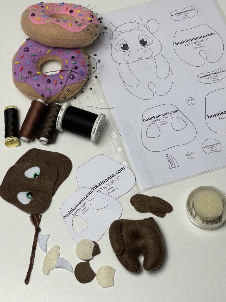 Felt calf sewing pattern pieces, thread, pincushions and partly stitched plush elements arranged for a handmade toy project
