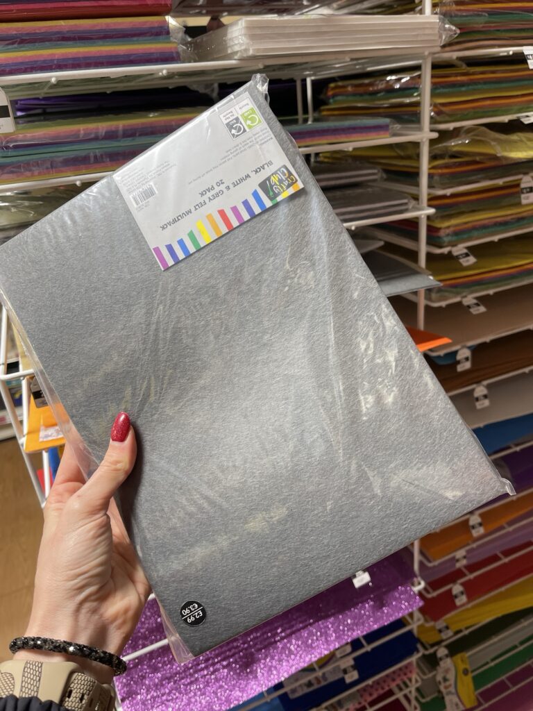 Hand holding stiff felt sheet in craft shop aisle with colourful felt stacks behind, choosing material for car organiser project