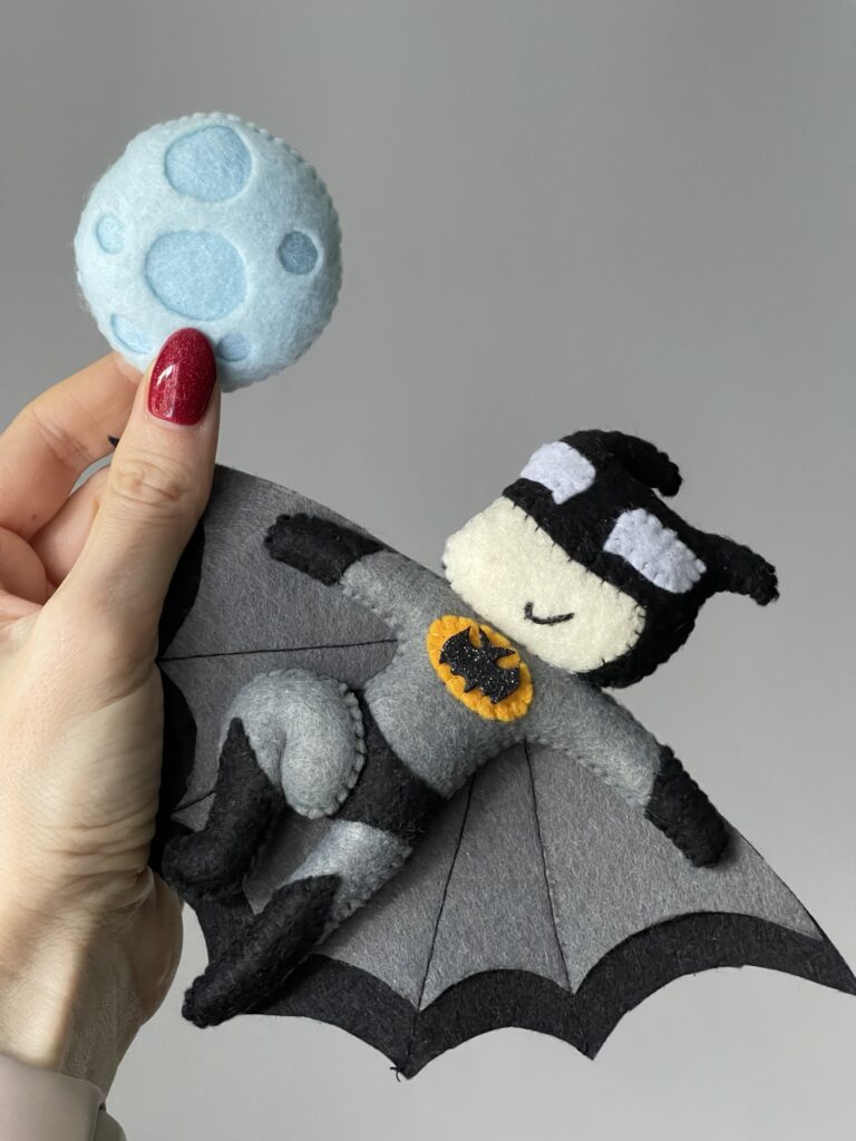 Hand holding finished felt Batman plush with stitched cape and moon detail, showing final toy result after pattern testing
