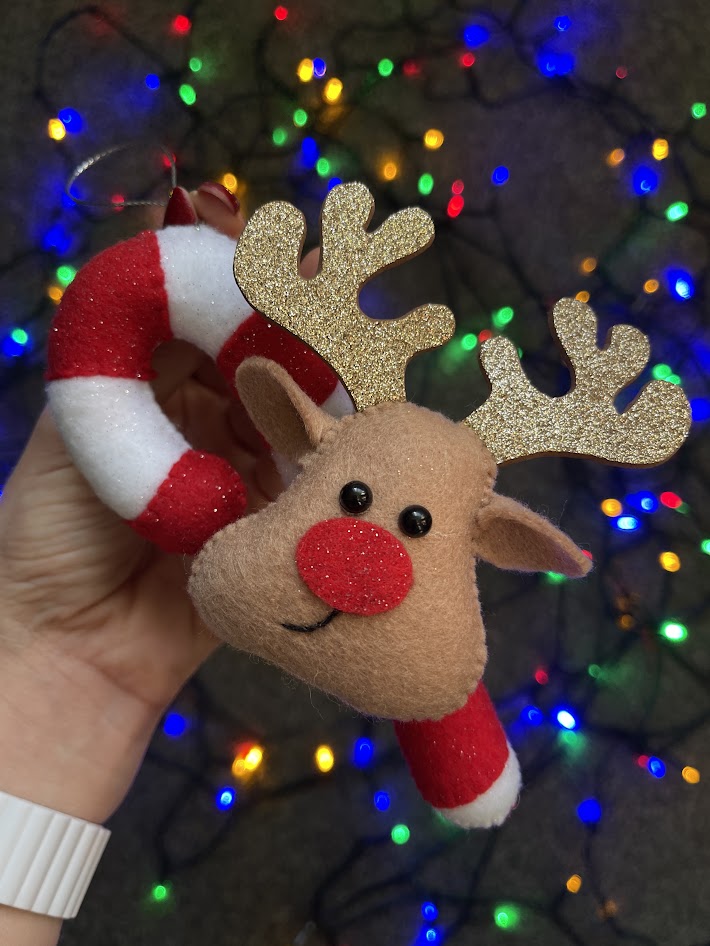 Glitter felt reindeer Christmas ornament with sparkly antlers and red nose held against festive fairy lights background