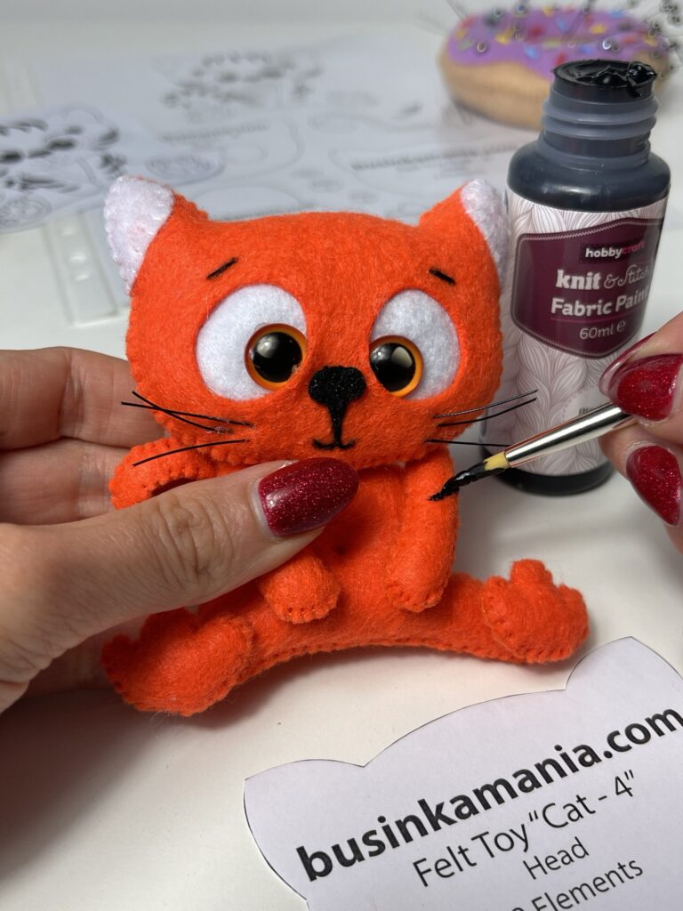 Hand painting black stripes onto a handmade felt tiger plushie using fabric paint and a thin brush during sewing assembly